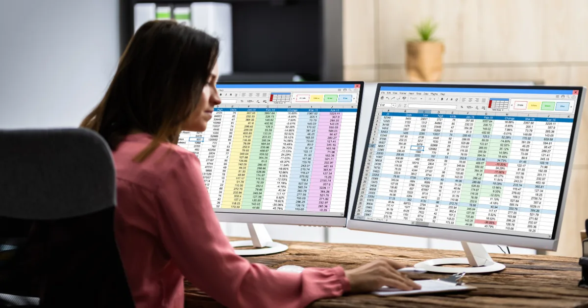 Business professional working on dual monitor setup with complex spreadsheets showing manual data entry and back-office operations