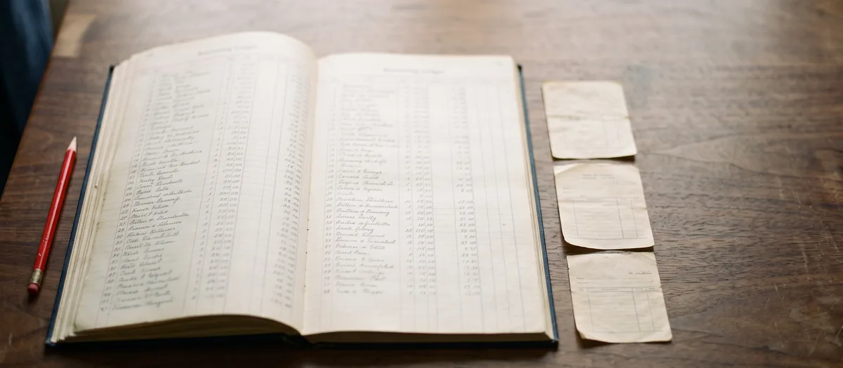 An open accounting ledger beside three paper receipts on a walnut table, with a red pencil.